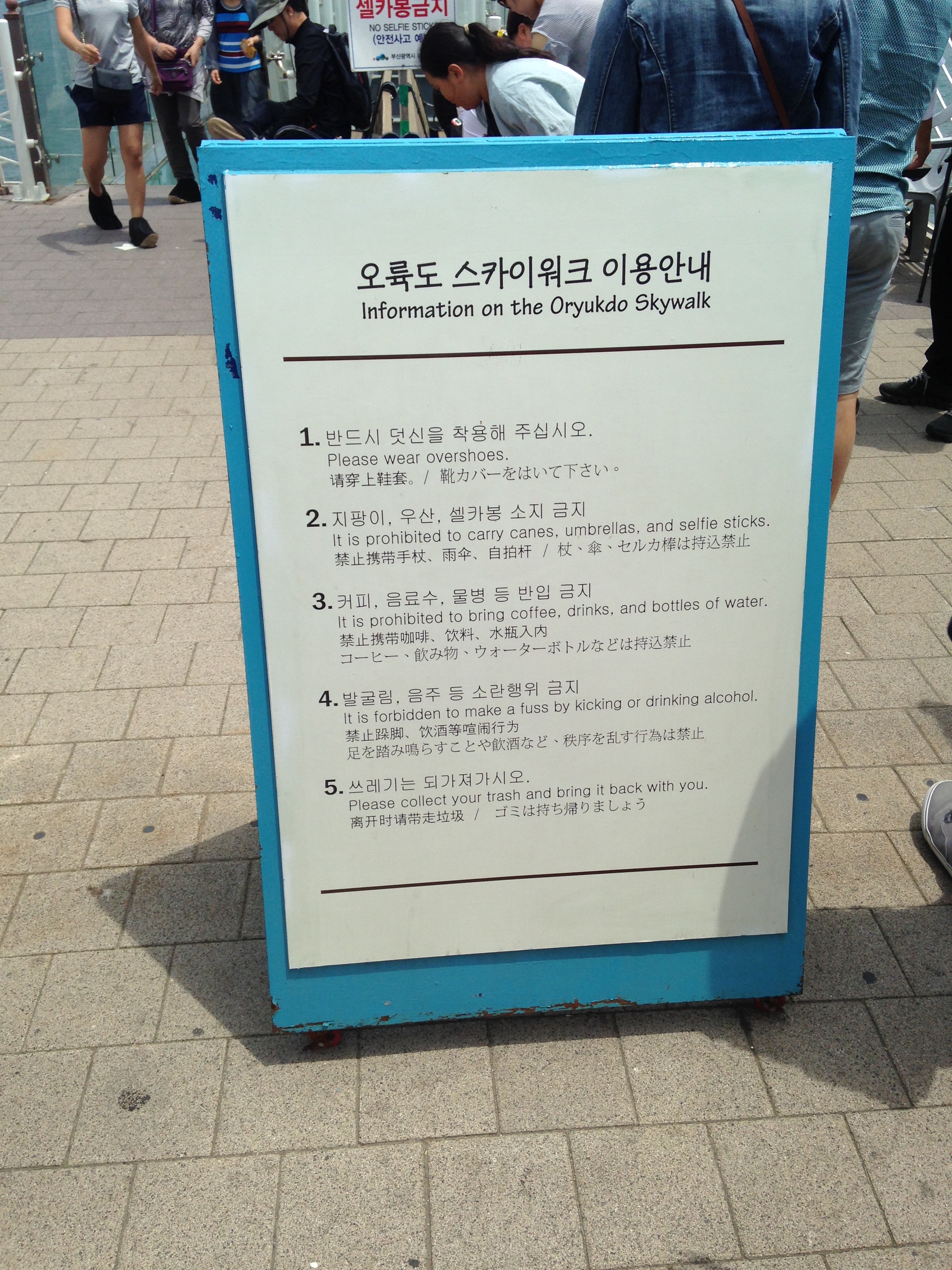 A sign on a blue easel in English, Korean, and several other Asian scripts. The English reads as follows: Information on the Oryukdo Skywalk / 1. Please wear overshoes. / 2. It is prohibited to carry canes, umbrellas, and selfie sticks. / 3. It is prohibited to bring coffee, drinks, and bottles of water. / 4. It is forbidden to make a fuss by kicking or drinking alcohol. /  5. Please collect your trash and bring it back with you.